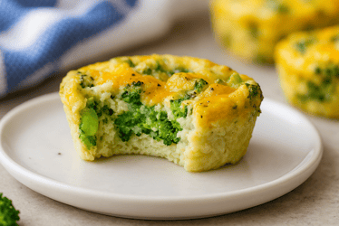 Muffin de huevo con brócoli y queso en un plato blanco al que se le ha quitado un bocado, mostrando el brócoli tierno y los huevos esponjosos con queso cheddar fundido por encima.